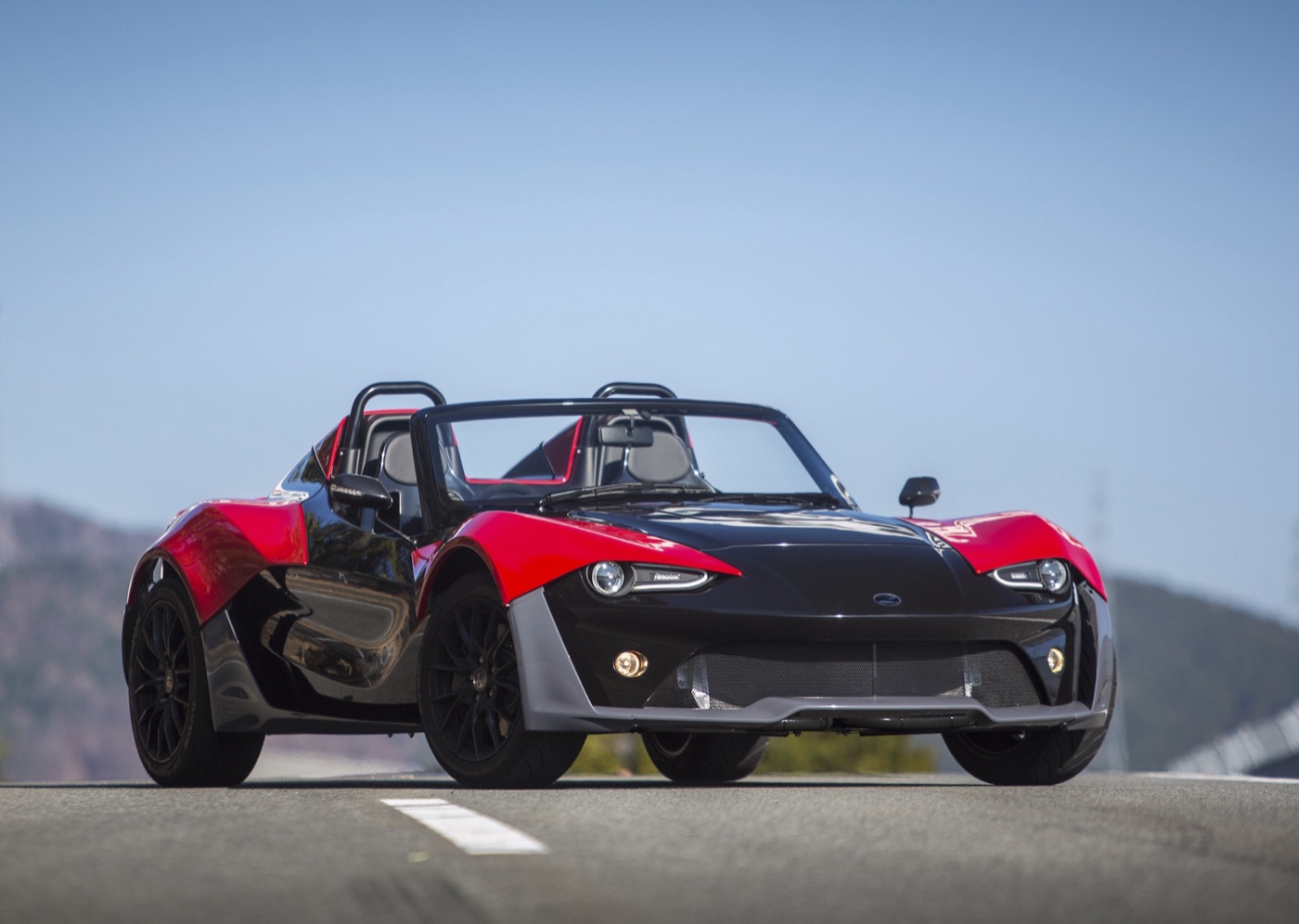 Red and black Zenos E10 roadster with twin roll hoops on an open road under a blue sky
