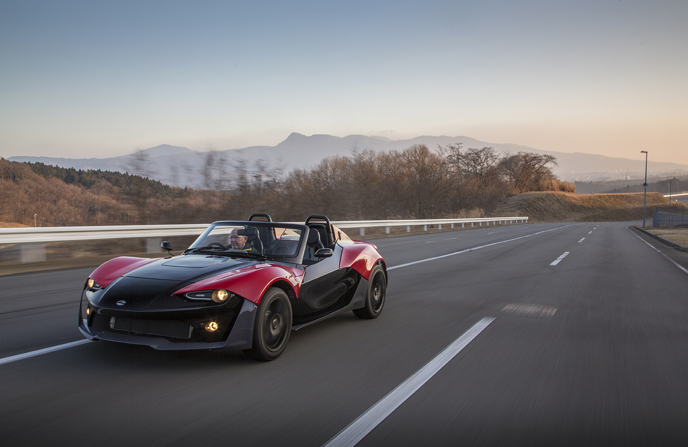 Red and black Zenos E10 driven on an empty road with mountains behind at dusk