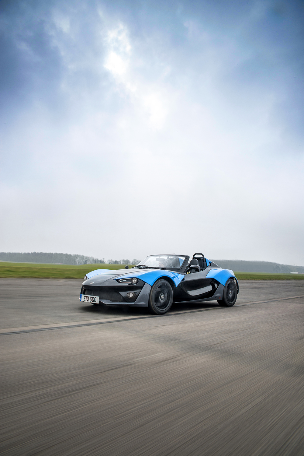 Blue and grey Zenos E10 S driving fast across an airfield under a grey sky
