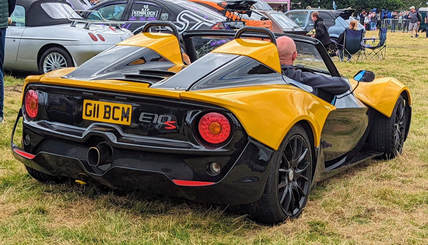 Yellow and black Zenos E10 S seen from behind, showing its round tail lights, centre exhaust and E10 S badge, at an outdoor car show