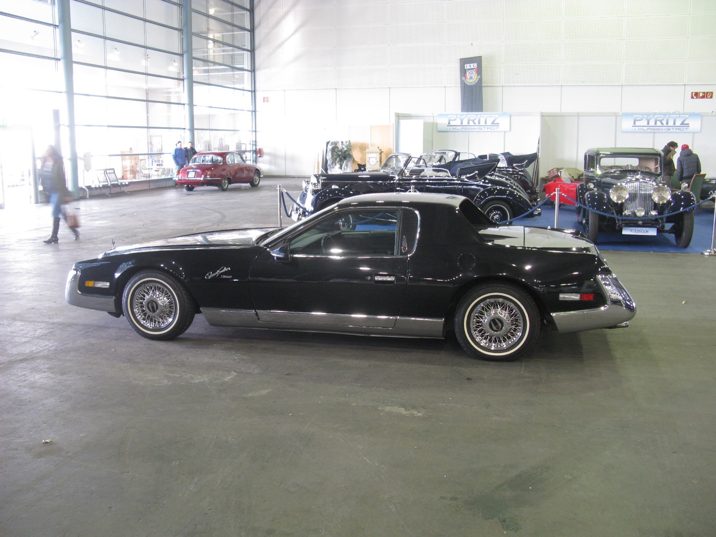 Black Zimmer QuickSilver coupé in side profile, with a long bonnet, a chrome sill and wire wheels, in an exhibition hall